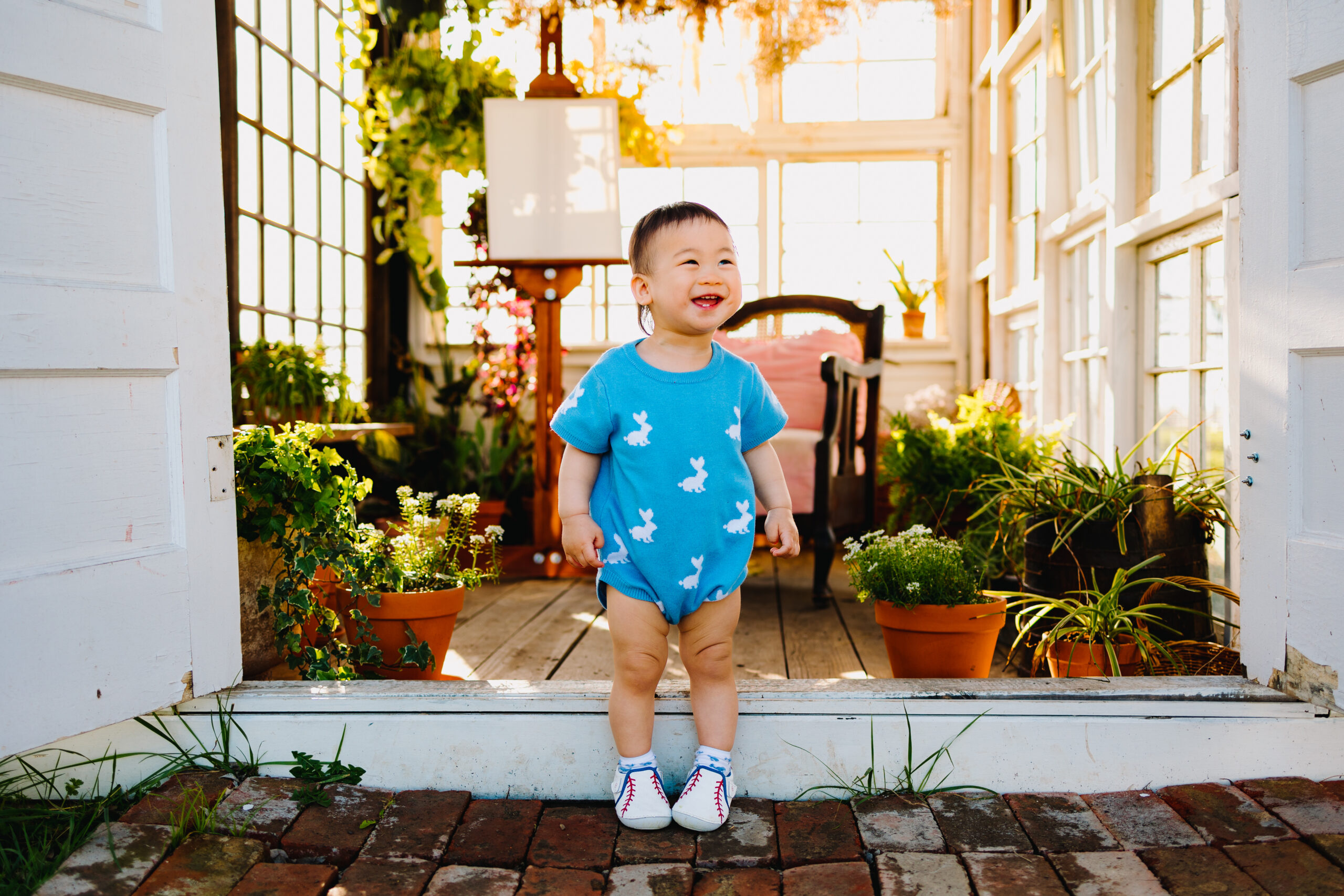a little boy in a blue romper with white bunnies on it stands in front of a white greenhouse full of plants and an artist's easel. He is smiling off to the side while looking at his parents.