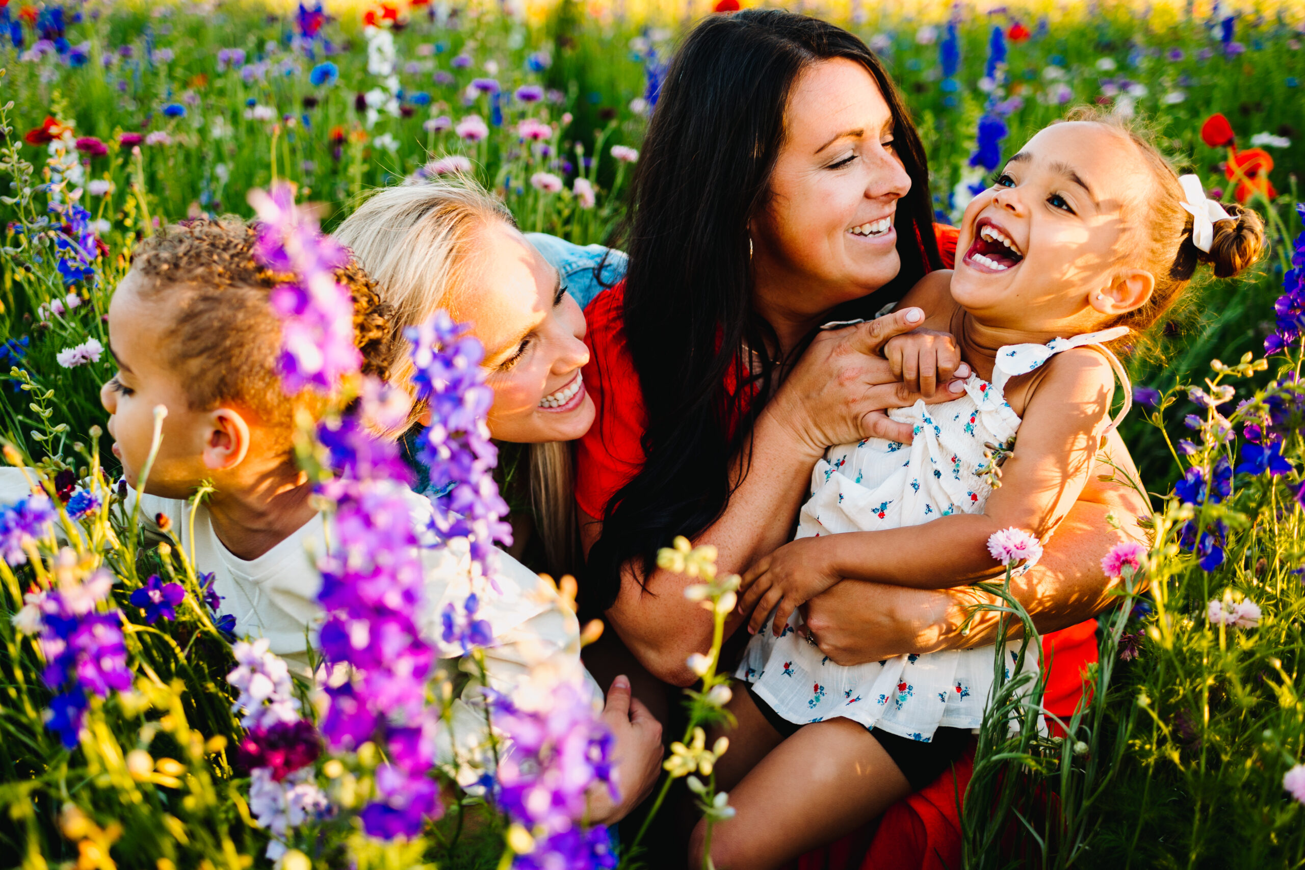 two moms cuddle with their twin toddlers in a wildflower field in richardson during a mini session. one mom is tickling her daughter while she laughs