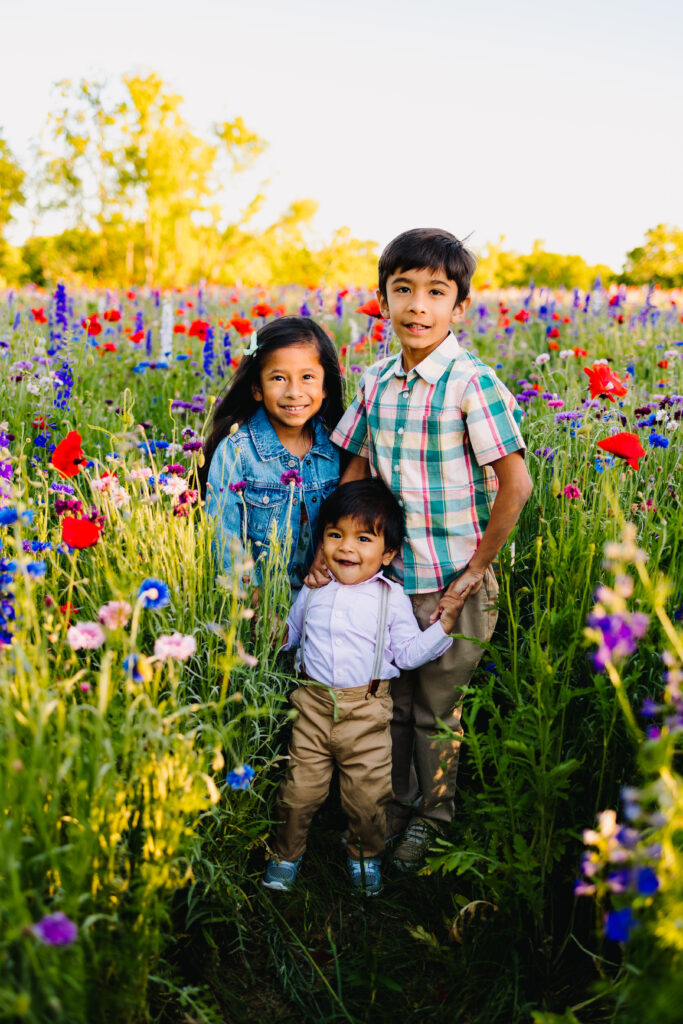 three siblings standing in a wildflower field during a mini session in dallas. All three kids are looking at the camera and smiling. dallas mini session photographer