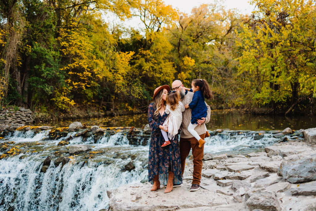 A family stands on the edge of a waterfall at Prairie Creek Park in Richardson, Texas. Beautiful fall foliage is behind them. The mom is holding her toddler daughter and the dad is holding their older daughter. Both parents are kissing the youngest daughter while the older girl tickles her dad.